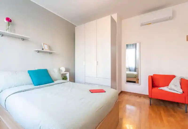 Dormitorio moderno y luminoso con cama grande, armario empotrado y un sillón naranja como punto focal. Textiles suaves y decoración minimalista ofrecen un ambiente acogedor, ideal para alquileres.