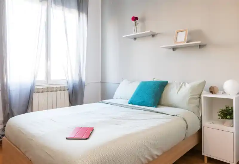 Dormitorio luminoso y bien decorado con cama doble, ropa de cama suave y un cojín turquesa; la luz natural entra por la ventana creando un ambiente tranquilo y acogedor.