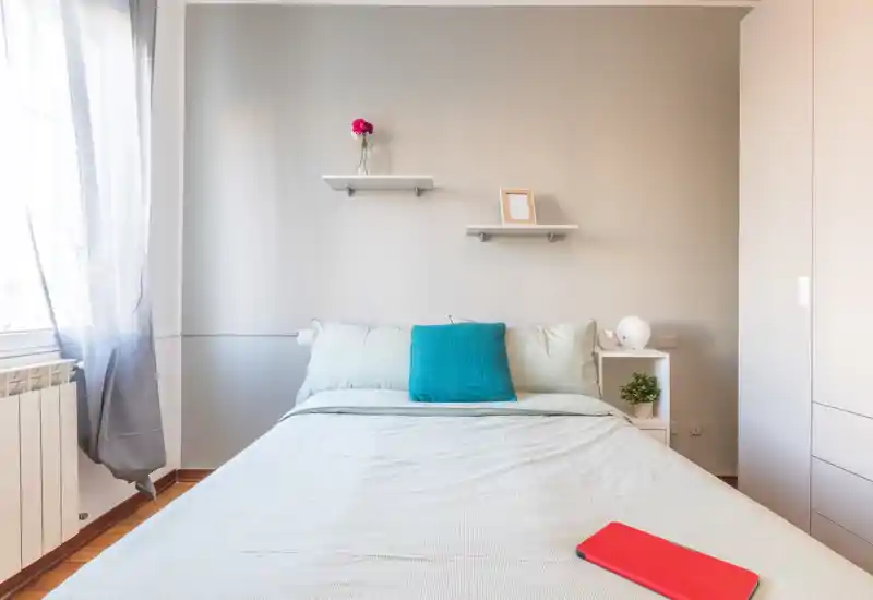 Dormitorio moderno y luminoso con cama doble bien hecha, cojín decorativo y estanterías sencillas: perfecto para anuncios de alquiler.