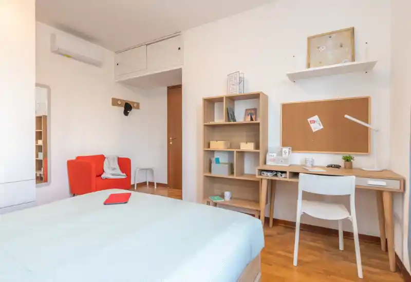 Dormitorio luminoso y bien presentado con la cama visible en primer plano, un escritorio con corcho, estanterías y un acogedor sillón rojo — ideal para una persona o pareja.