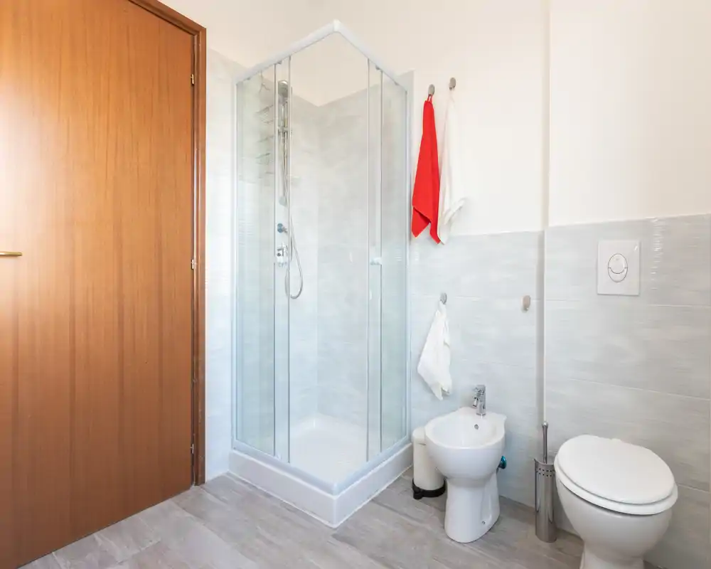 Baño moderno y limpio con ducha angular de cristal, bidé y váter, azulejos neutros y luz natural brillante.