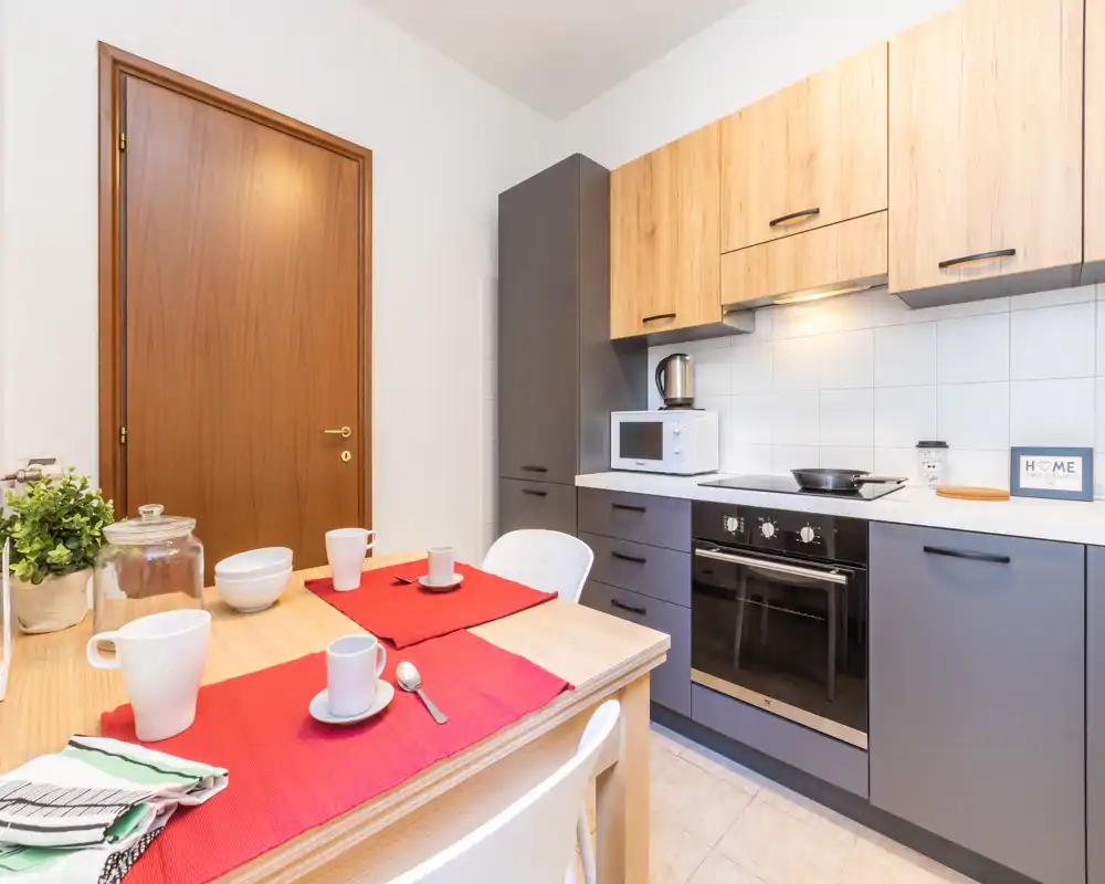 Cocina moderna y luminosa con horno empotrado, microondas y una mesa pequeña con manteles rojos — ideal para anuncios de apartamentos compactos.