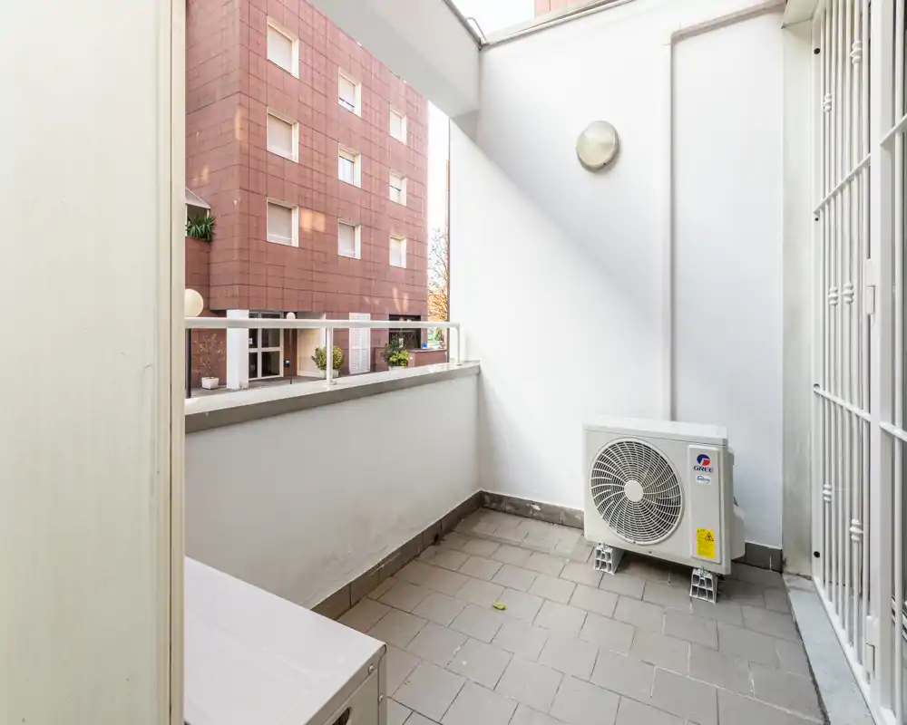 Pequeño balcón cubierto con suelo de baldosas, vista parcial del edificio vecino y unidad exterior del aire acondicionado. Espacio luminoso y resguardado, apto para plantas o un pequeño rincón de descanso.