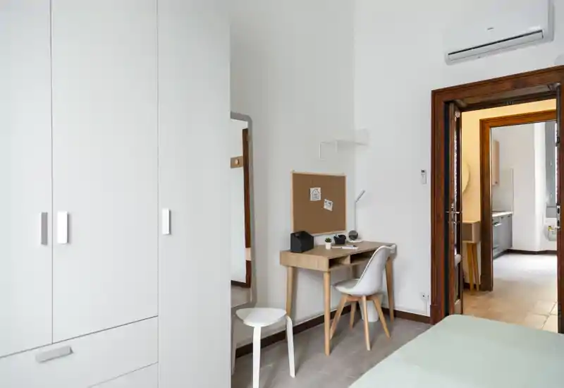 Camera da letto luminosa e minimal con armadio, piccolo scrittoio in legno e letto parzialmente visibile al bordo dell'inquadratura. Stile pulito e moderno con pareti neutre e luce naturale.