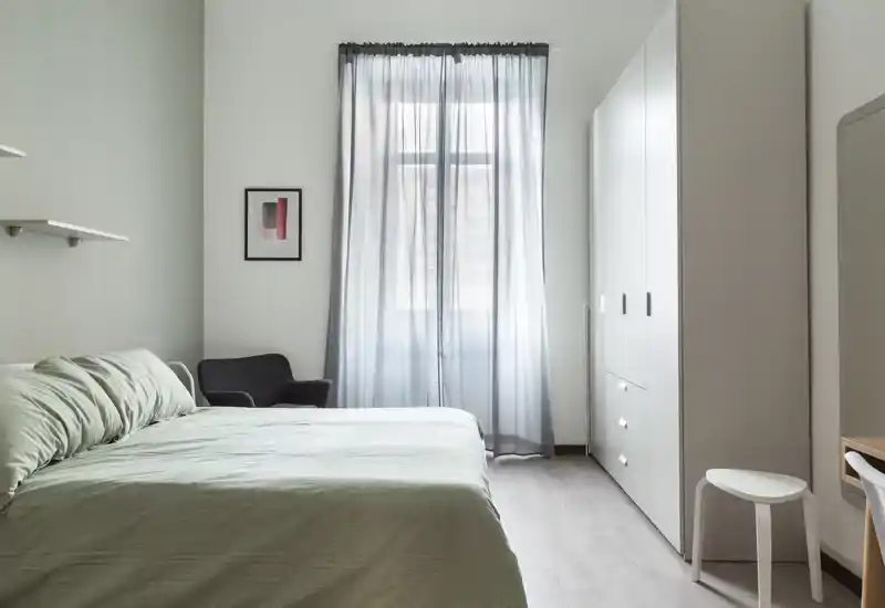 Camera da letto minimal e luminosa con letto matrimoniale ben rifatto, lenzuola verde tenue, un armadio e una poltrona accanto a una grande finestra che offre luce naturale.