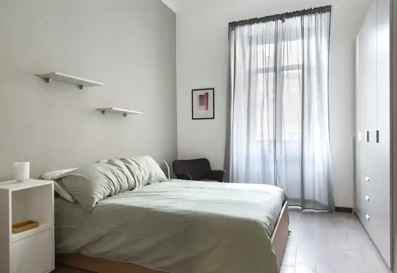 Camera da letto luminosa e minimal con letto matrimoniale in lenzuola verde tenue, comodino a cubo, poltrona e armadio spazioso vicino a una finestra con tenda leggera.
