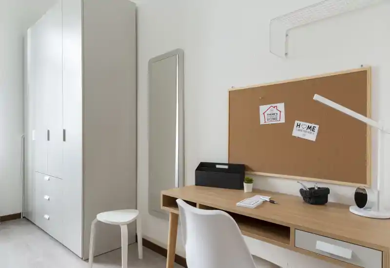 Angolo studio luminoso e minimale con scrivania in legno, bacheca in sughero, sedia bianca e guardaroba — ideale per studiare o lavorare da casa.