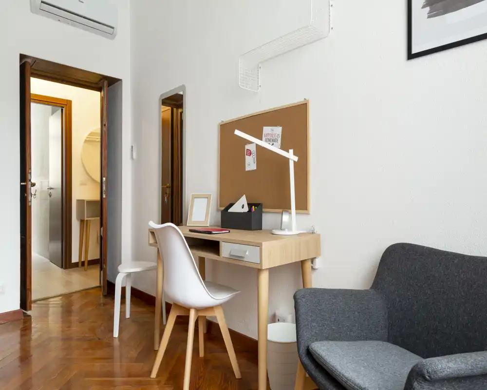 Angolo living accogliente con poltrona moderna, scrivania in legno e bacheca: ideale per rilassarsi o lavorare in uno spazio compatto.