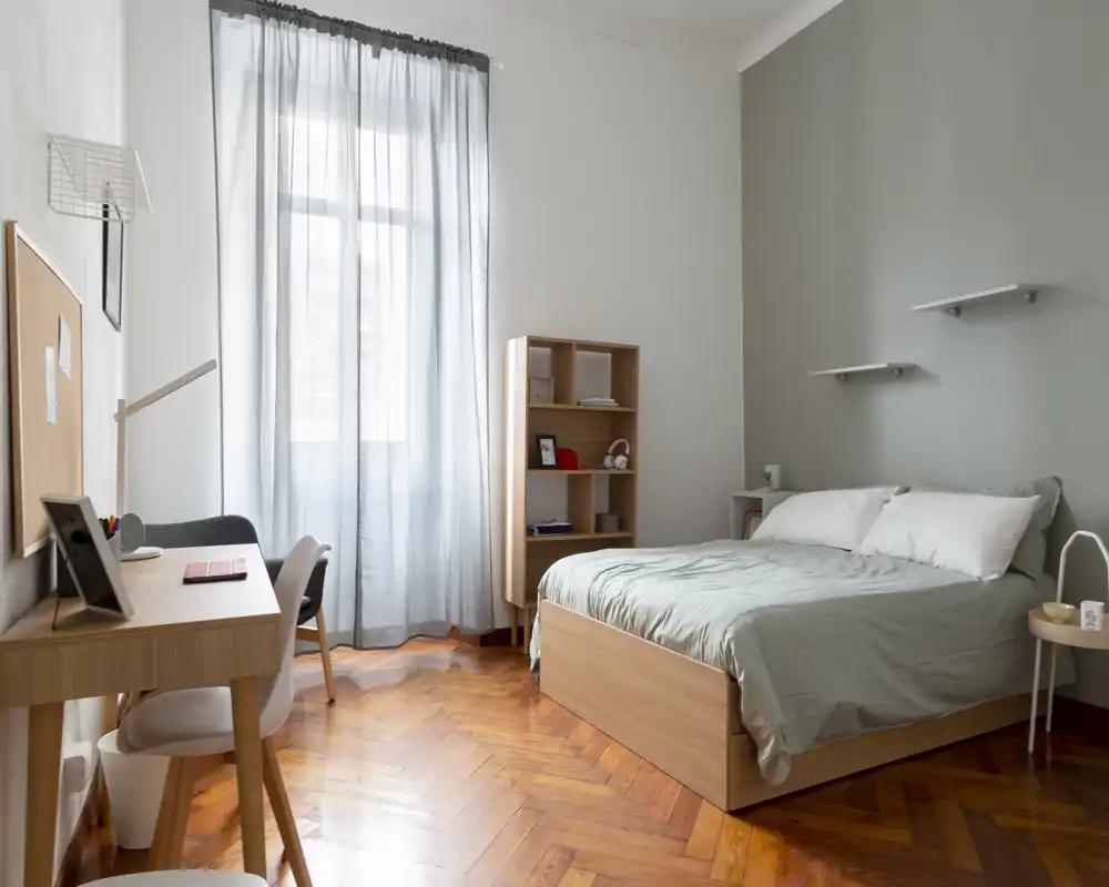 Camera da letto luminosa e minimale con letto matrimoniale, comodino e una piccola scrivania vicino a una finestra alta; parquet in legno caldo e tessuti grigio-verdi creano un'atmosfera calma e ariosa.