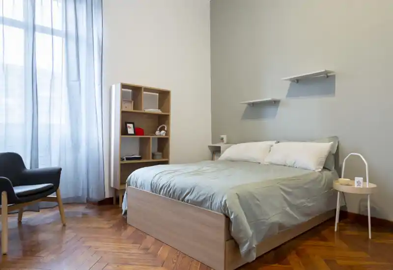 Camera da letto luminosa e ordinata con letto matrimoniale, lenzuola chiare e un piccolo comodino; la luce naturale filtrata dalle tende leggere e il pavimento in legno caldo creano atmosfera.