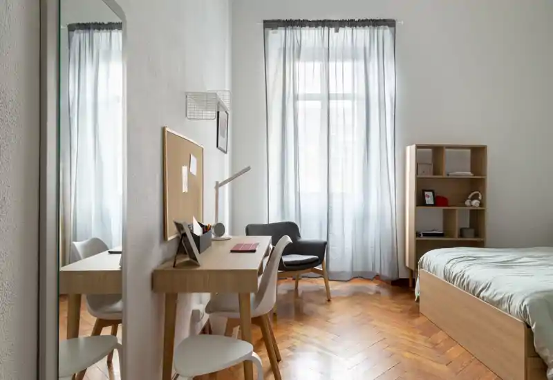Camera da letto luminosa e minimal con letto in legno, scrivania a muro, poltroncina e scaffale — luce naturale filtrata dalle tende leggere crea un'area notte e studio tranquilla.