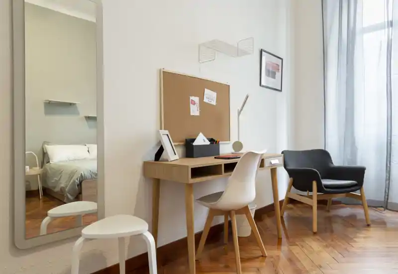 Angolo camera luminoso e ben arredato con scrivania e poltrona. Il letto è visibile attraverso la porta e nello specchio; la luce naturale filtrata dalle tende crea un angolo lavoro accogliente.