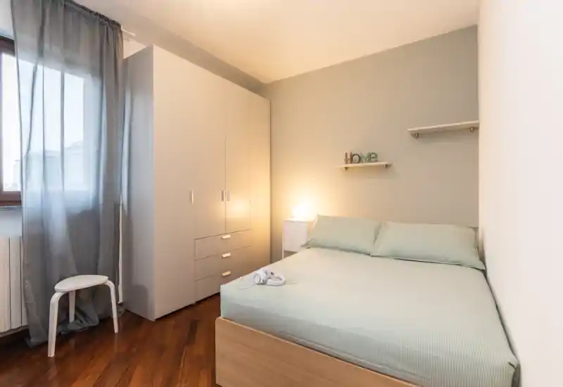 Bright, compact bedroom with a double bed, bedside table and wardrobe; soft green linens and warm lighting create a calm, welcoming atmosphere.