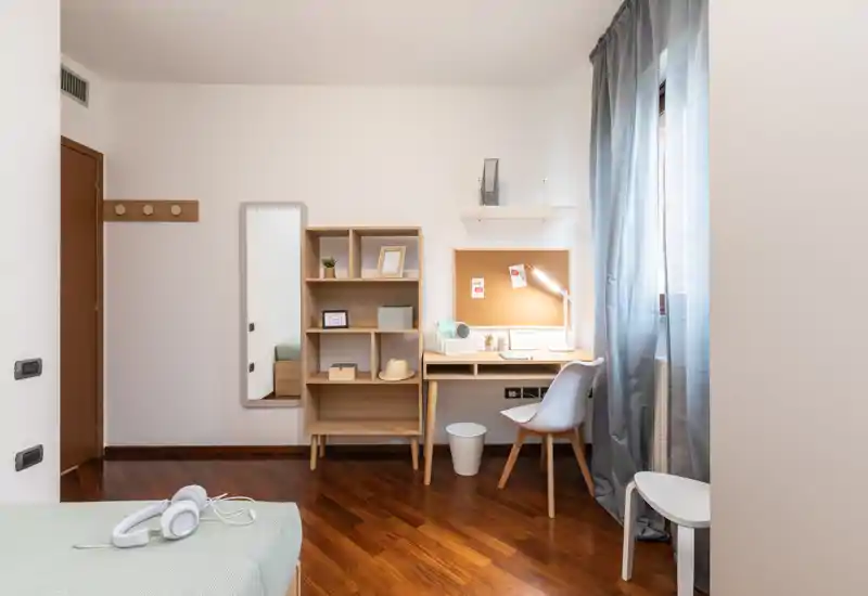 Well-lit bedroom corner with a single bed partially visible, a tidy wooden desk and shelving unit by the window — ideal for a student or remote worker.