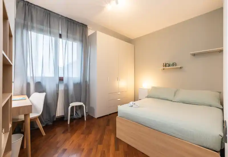 Bright, well-staged bedroom with a double bed, wardrobe and small desk by the window; warm lamp lighting and pale green bedding create a calm atmosphere.