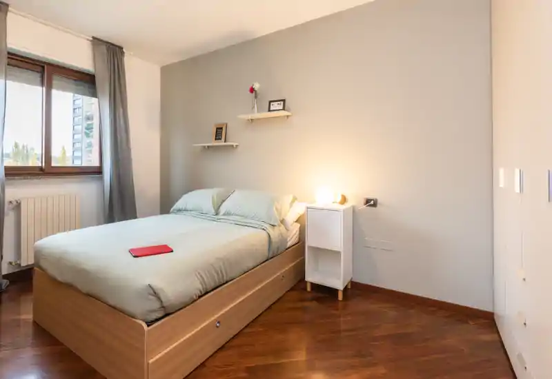Camera da letto luminosa e ben allestita con letto matrimoniale, biancheria morbida e lampada da comodino. La finestra ampia porta luce naturale e il pavimento in legno aggiunge calore — perfetta per annunci.