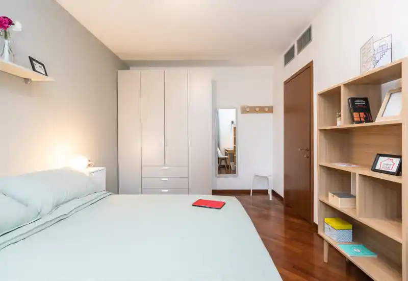 Camera da letto luminosa e moderna con letto matrimoniale ben rifatto, armadio, specchio a figura intera e scaffalatura in legno — arredata con gusto e ordine.