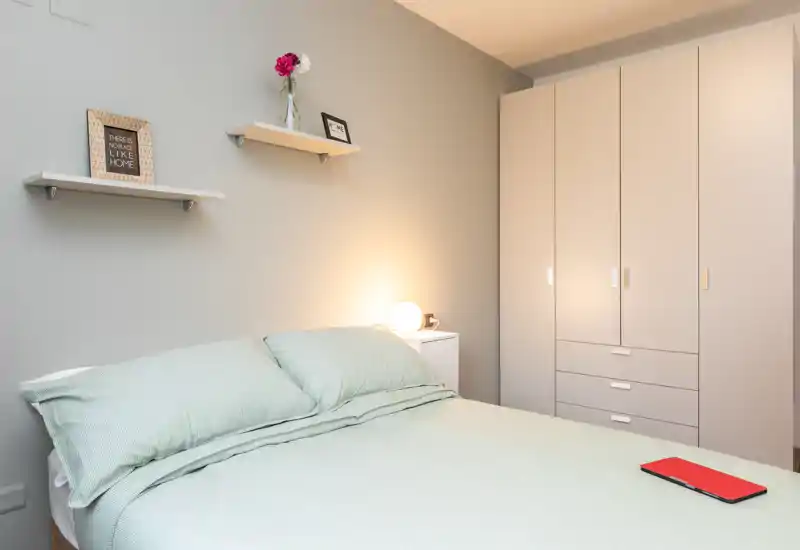 Camera da letto luminosa e minimal con letto matrimoniale rifatto, lampada da comodino e armadio a muro — linee pulite e biancheria pastello creano un ambiente rilassante.