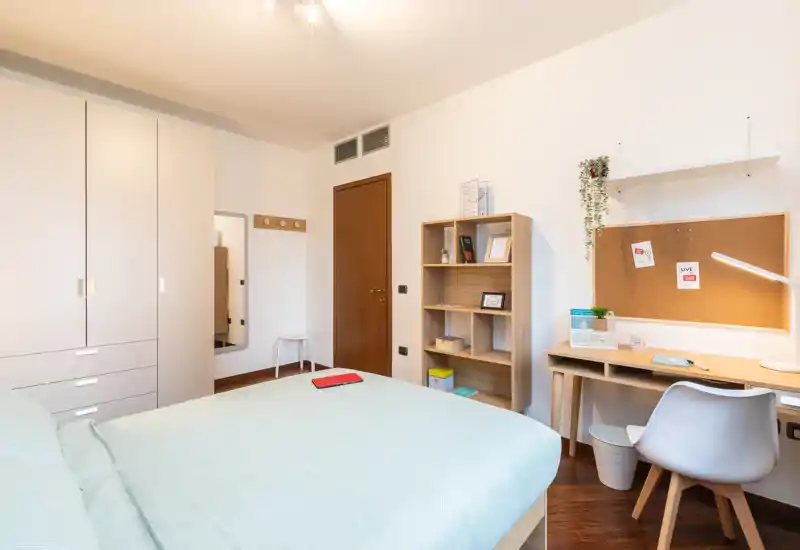 Camera da letto luminosa e moderna con letto ampio, armadio, scrivania e scaffalatura — ideale per un soggiorno confortevole con luce naturale e arredo minimal.
