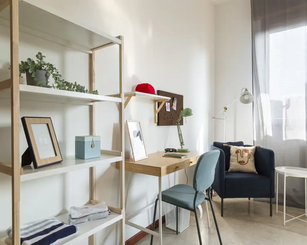 Rincón de estar luminoso y acogedor con un sillón cómodo, un pequeño escritorio y estanterías — ideal para relajarse o trabajar junto a la ventana.