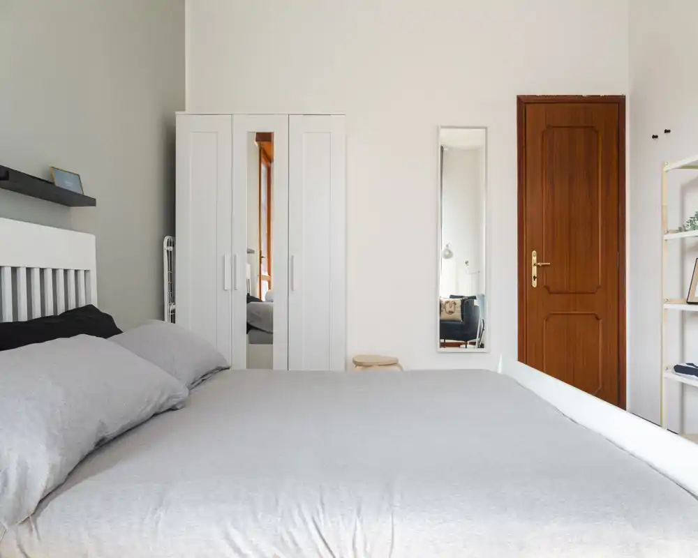 Dormitorio luminoso y minimalista con una cama grande en ropa de cama neutra, un armario blanco con puerta espejo y un espejo de cuerpo entero junto a la puerta de madera — limpio y aireado.