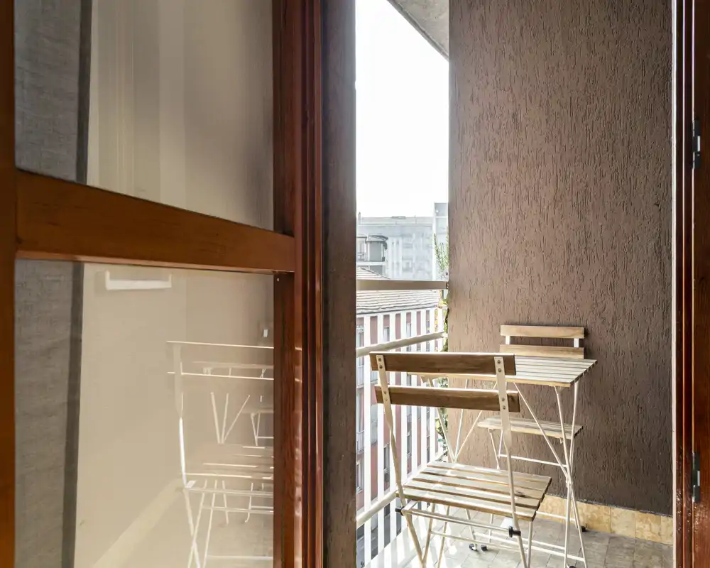 Pequeño balcón amueblado con mesa y sillas plegables de madera, suelo de baldosas y barandilla metálica; buena luz natural y vista parcial de la ciudad.