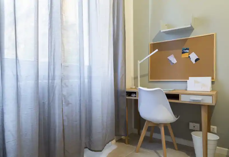 Rincón de trabajo ordenado con escritorio de madera, silla blanca, lámpara y corcho; las cortinas tamizan la luz natural para un área de estudio tranquila.