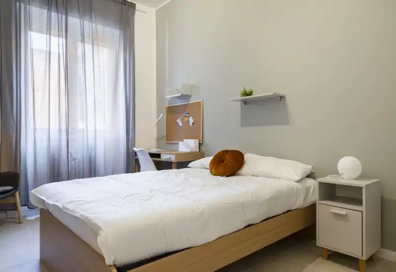 Dormitorio moderno y luminoso con cama de madera, ropa de cama blanca y una mesita; un escritorio junto a la ventana recibe luz natural suave filtrada por las cortinas.