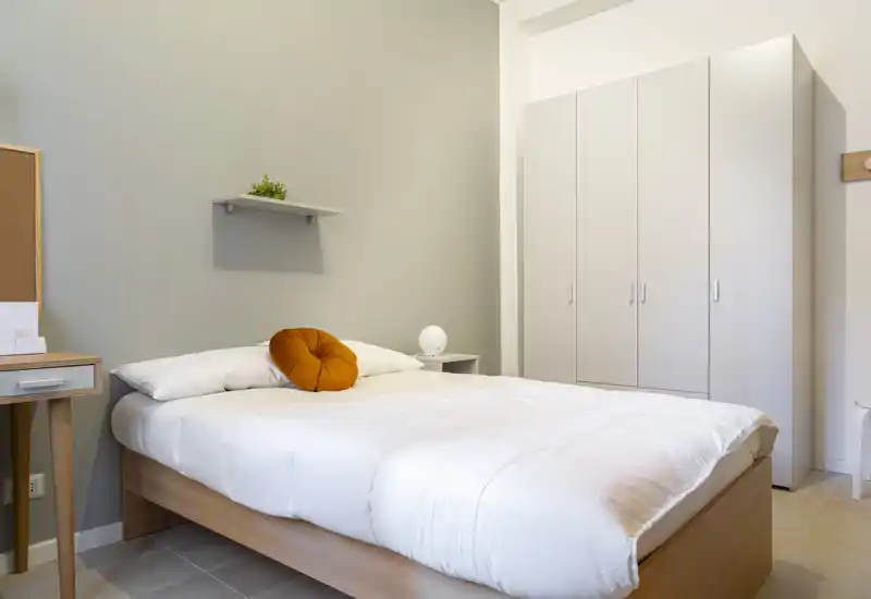 Dormitorio moderno y luminoso con cama completa, ropa blanca y un cojín de acento cálido; armario y mesita de noche sencillos para un ambiente minimalista.