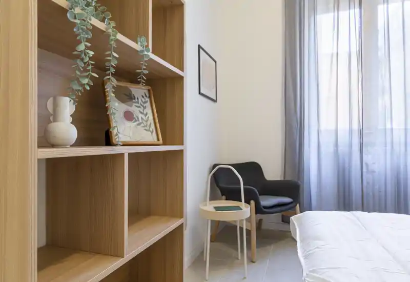 Rincón de dormitorio luminoso y moderno con cama visible, butaca acogedora y estantería decorativa; cortinas traslúcidas dejan entrar luz natural suave.