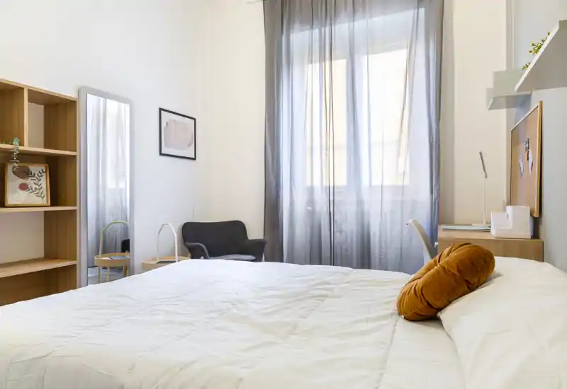 Dormitorio luminoso y moderno con una cama grande en primer plano, ropa de cama blanca y un cojín redondo decorativo; cortinas translúcidas filtran la luz natural y un pequeño escritorio y sillón completan la habitación.