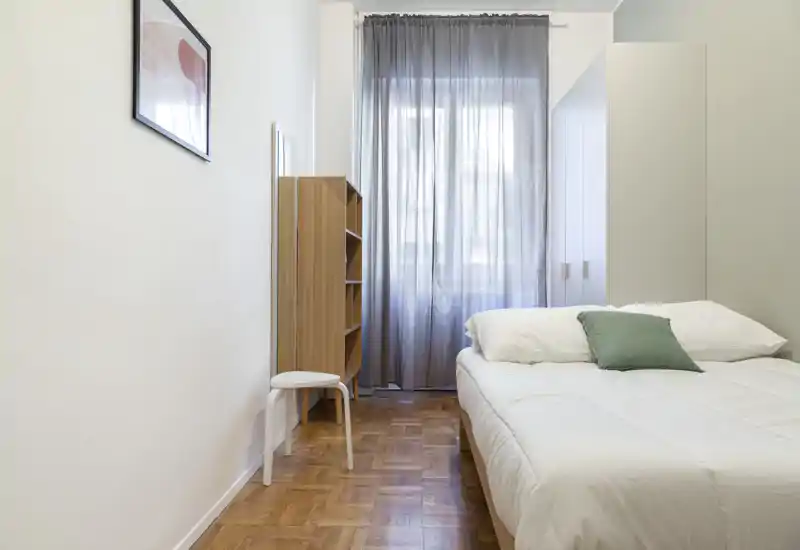 Bright, minimalist bedroom with a neatly made double bed, soft linens and a green accent cushion; sheer curtains let in natural light and there is a white wardrobe and wooden shelving for storage.