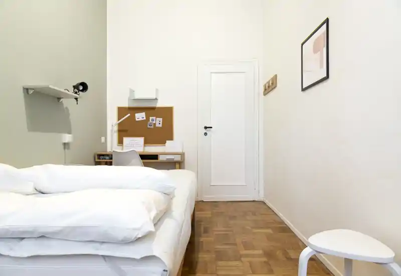 Minimal, bright bedroom with a large bed, wooden parquet floor and a small desk; neutral tones and simple decor create a calm, inviting space.