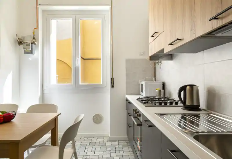 Modern compact kitchen with a gas hob, stainless sink, kettle and microwave; a wooden dining table and chairs by a bright window create a cozy dining corner.