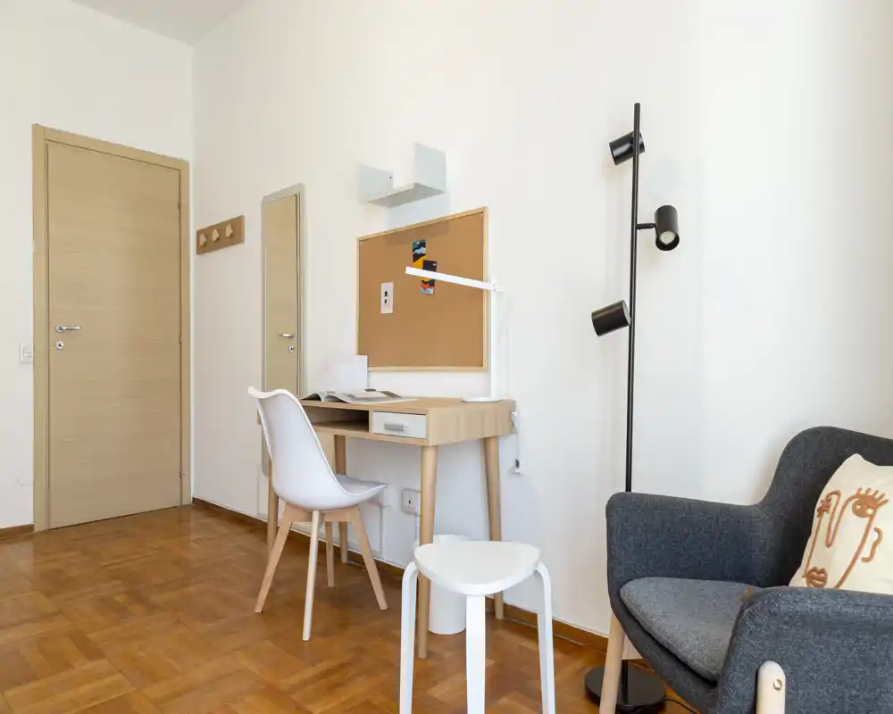 Zona de estar luminosa y ordenada con un pequeño escritorio de madera, silla y un sillón cómodo; decoración minimalista y suelo de parquet cálido que crean un rincón de trabajo acogedor.