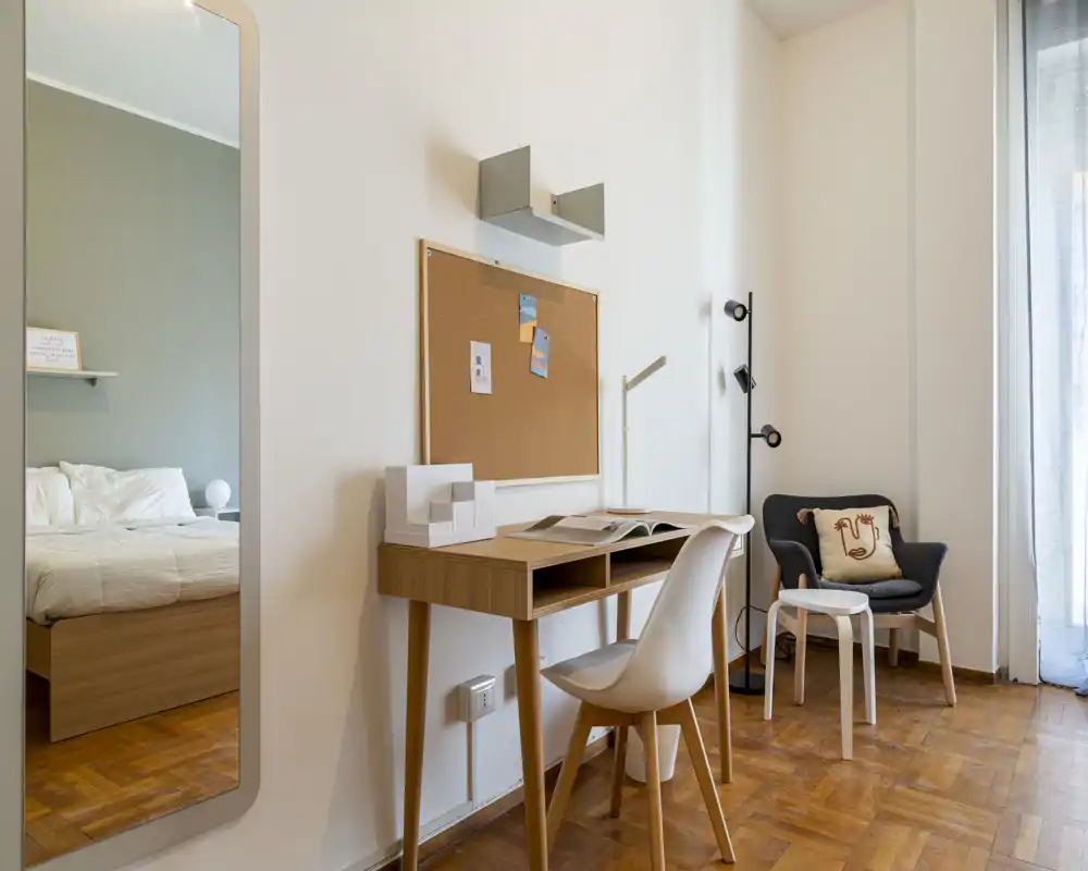 Rincón de dormitorio con estilo: cama de madera visible a la izquierda (parcialmente), escritorio compacto con silla, butaca acogedora y luz natural suave desde el balcón — perfecto para una persona o estudiante.