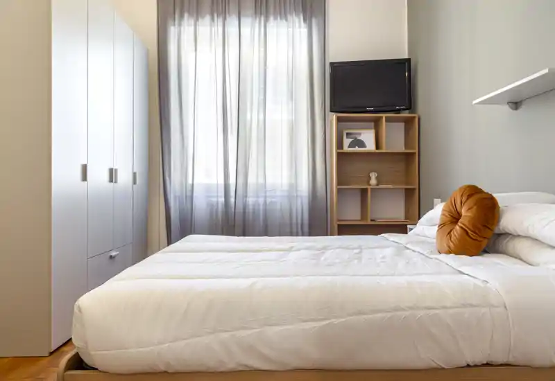 Dormitorio luminoso y ordenado con cama doble cómoda, ropa de cama suave y un cojín redondo decorativo; armario y estantería para almacenamiento y cortinas que tamizan la luz natural.
