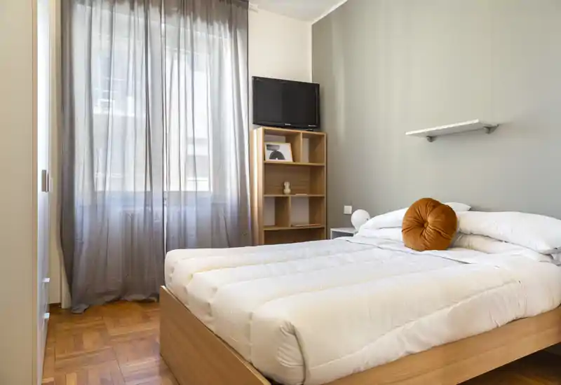Dormitorio luminoso y ordenado con cama doble, ropa de cama blanca y un cojín redondo decorativo; la luz natural entra a través de las cortinas translúcidas y el suelo de madera aporta calidez.