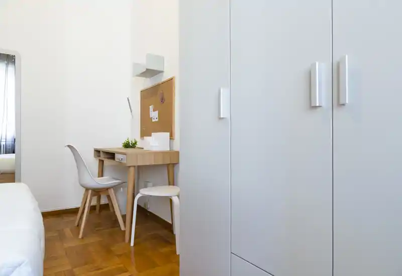 Bright, modern bedroom corner with a white bed, wooden parquet floor and a small study desk with chair — a clean, minimalist space ideal for short-term stays.