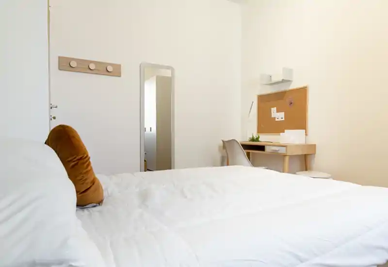 Dormitorio luminoso y minimalista con una gran cama blanca, cojín de color cálido y un pequeño escritorio de madera con corcho — ordenado, moderno y acogedor.
