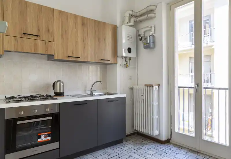 Compact modern kitchen with wood upper cabinets, dark lower units, gas hob and oven. Large glass door provides abundant natural light and access to a small balcony.