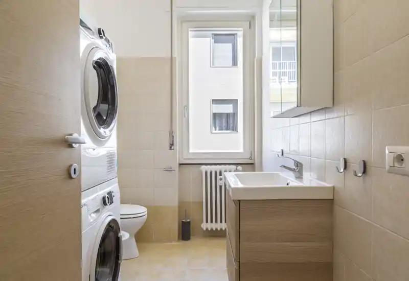 Bright, modern bathroom with a sink, vanity cabinet, toilet and stacked washer-dryer; neutral beige tiles and natural window light.