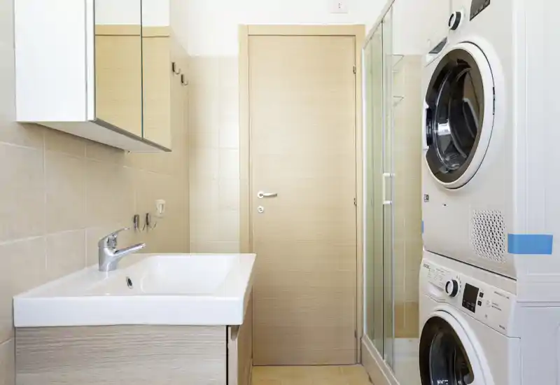 Bright, modern bathroom with a sink, mirror cabinet and stacked washer and dryer next to a glass shower cubicle. Neutral tiles and light wood finishes give a clean, functional look ideal for rental listings.