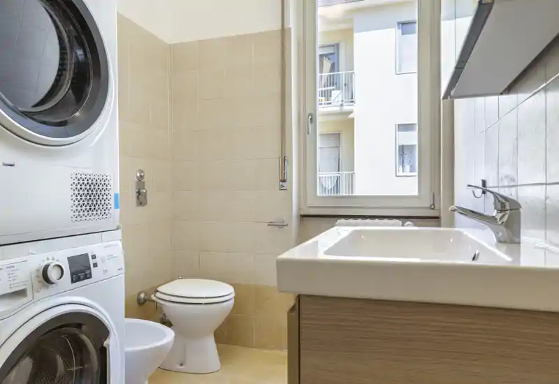 Compact, well-lit bathroom with a modern sink, toilet and a stacked washer-dryer unit; light tiles and a large window provide good natural light.