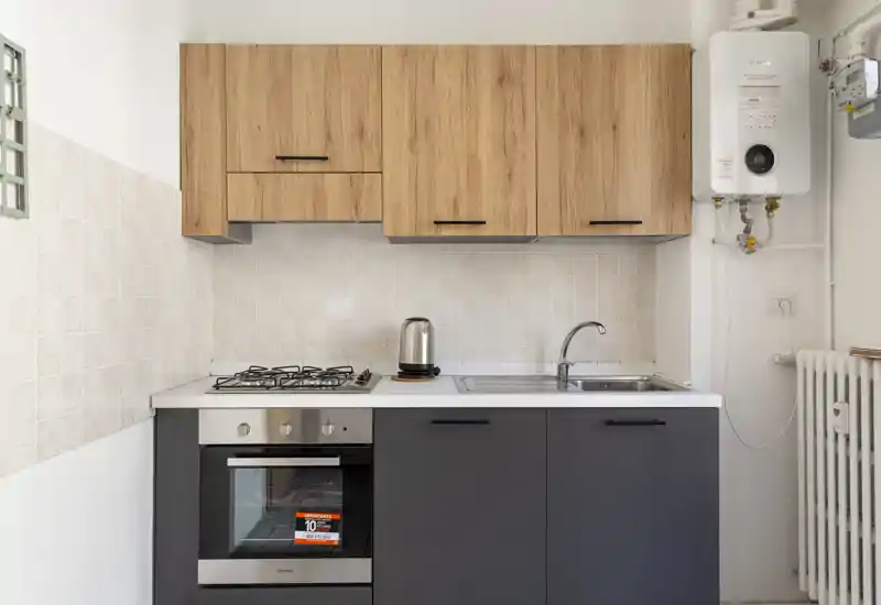 Compact modern kitchenette with wood-effect wall cabinets, dark base units, built-in oven, gas hob and stainless sink — bright and neatly finished, ideal for a rental listing.