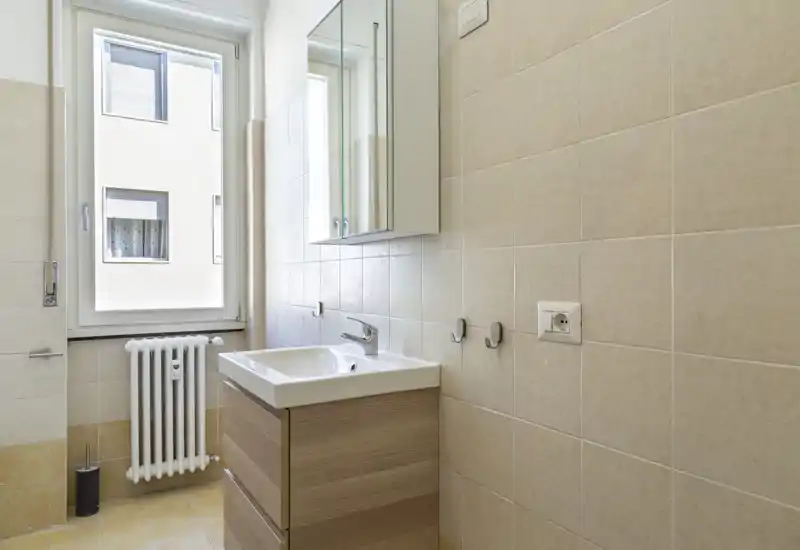 Bright, compact bathroom with a wall-mounted mirror cabinet, single sink on a wooden vanity and tiled walls; natural light from a large window.