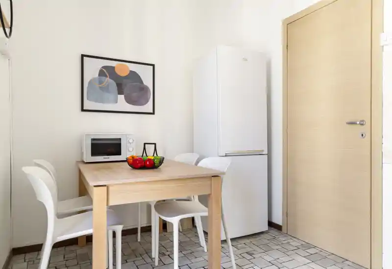 Compact kitchen/dining corner with a wooden table, four white chairs, refrigerator and microwave. Bright, tidy space styled with a fruit bowl and wall art, ideal for small apartments.