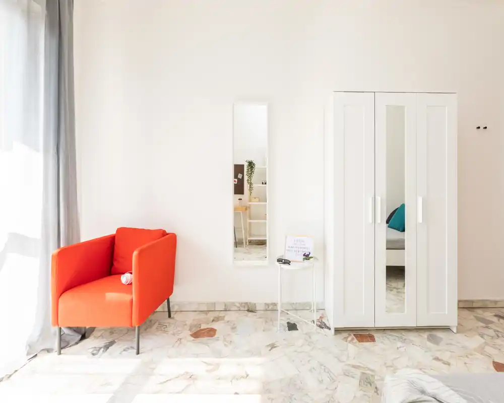 Rincón de dormitorio minimalista y luminoso con un armario blanco, espejo de cuerpo entero y un sillón naranja vivo. La luz natural y el suelo de mármol crean un ambiente fresco y aireado, ideal para anuncios.