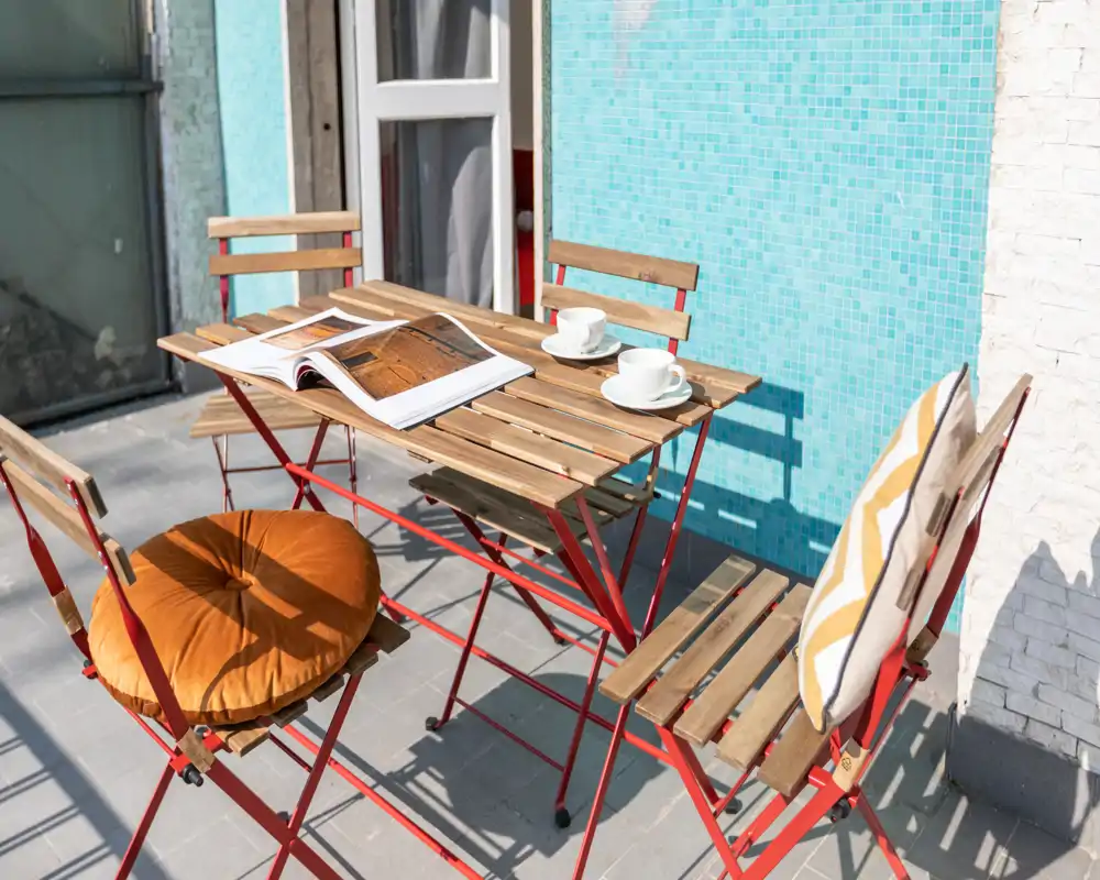 Pequeña terraza soleada con mesa bistró de madera y cuatro sillas plegables, cojines decorativos y dos tazas — un rincón luminoso y acogedor para el café matutino.