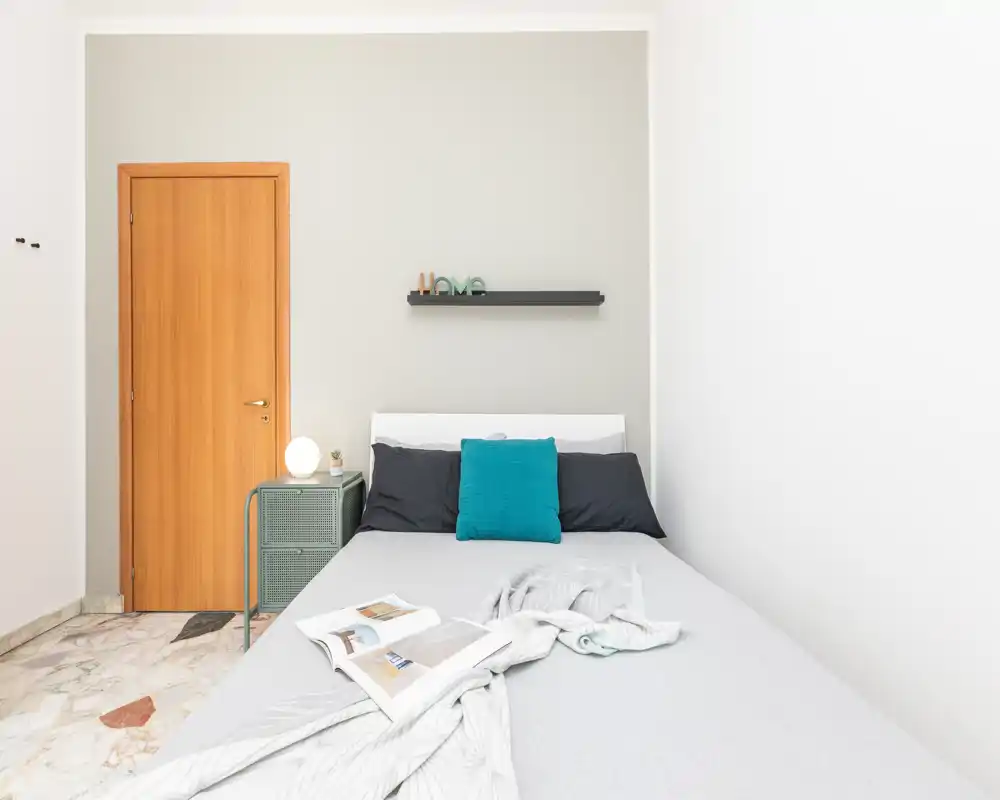 Dormitorio luminoso y minimalista con cama doble bien hecha, cojín turquesa y una mesita con lámpara — ideal para una estancia tranquila.